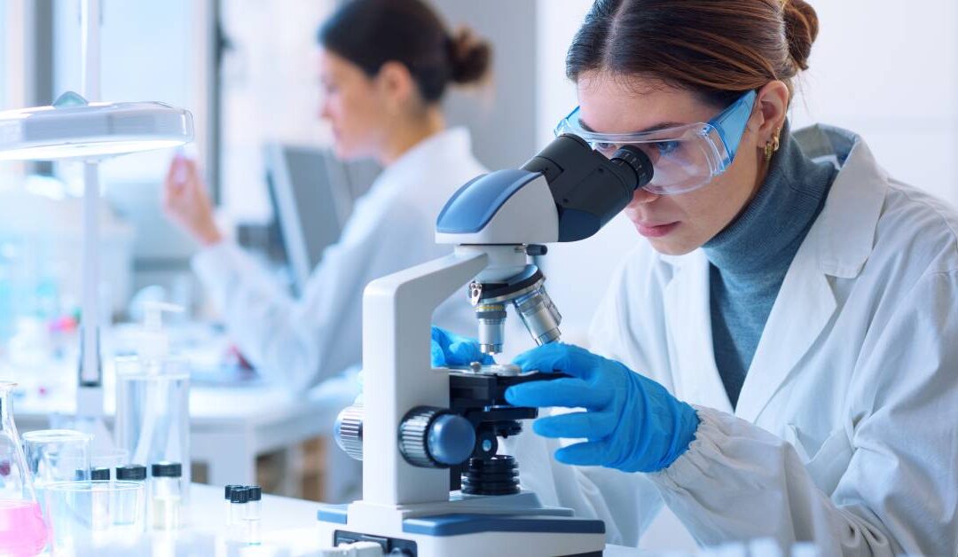 Two women scientists are in a research lab. One is looking through a microscope, and the other is examining a tube.