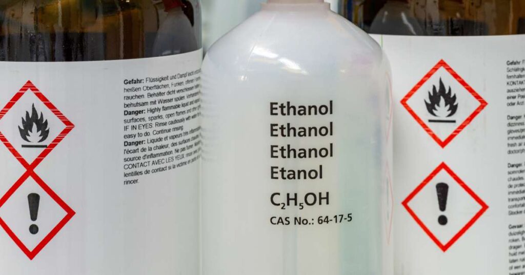 A bottle of ethanol on a table with other bottles. The word “Ethanol” is written four times and has its chemical formula.