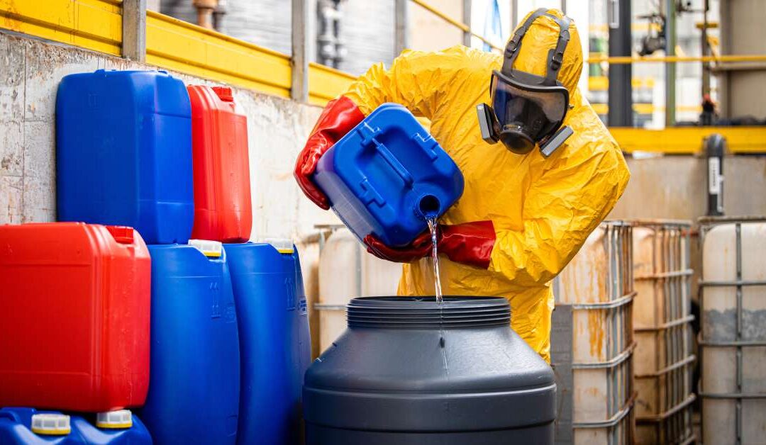 A worker wearing a hazmat suit is walking with canisters of solvents. There are several solvent canisters near him.