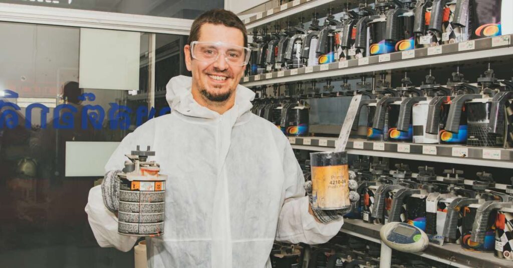 A professional wearing protective goggles and a suit holding cans of paint and paint thinner. He’s next to a shelf with paint.