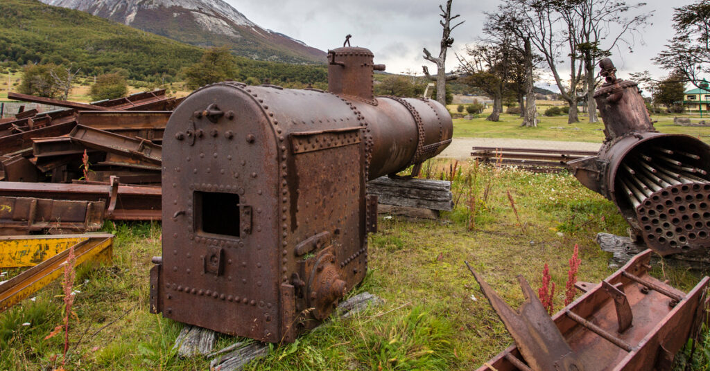 Salvageable train parts for restoration projects sit in a junkyard near trees and a large mountain.