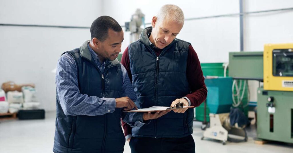 Two workers look at paperwork in a warehouse. One worker is holding a clipboard, and the other is pointing at information.