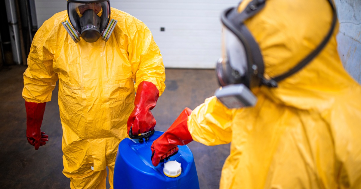 Two workers are wearing yellow protective suits and black gas masks while handling chemicals in a blue container.