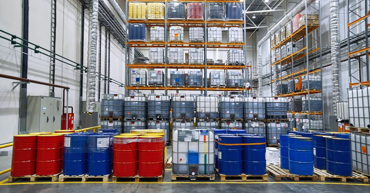 A factory with barrels of solvents on pallets stored on the ground and others stacked vertically on shelves.