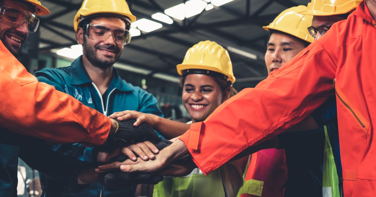 Factory workers wear yellow helmets, coveralls, and goggles as they put their hands together in a circle.