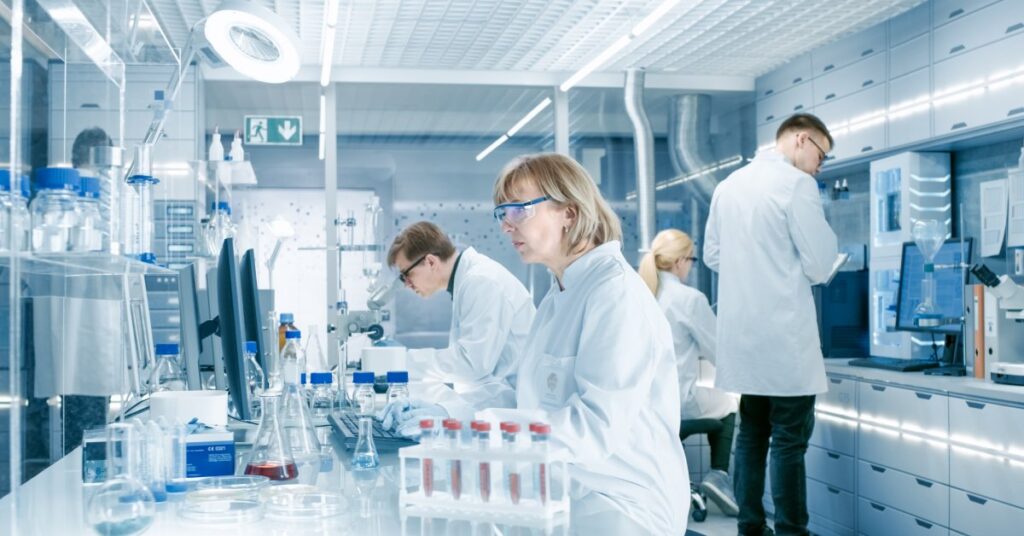 A group of scientists working in a pharmaceutical lab. They're all wearing white lab coats and protective eyewear.