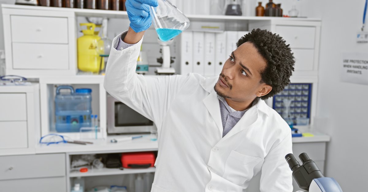 An African American man inspecting a blue chemical in a glass beaker. He's sitting at a lab station near a microscope.