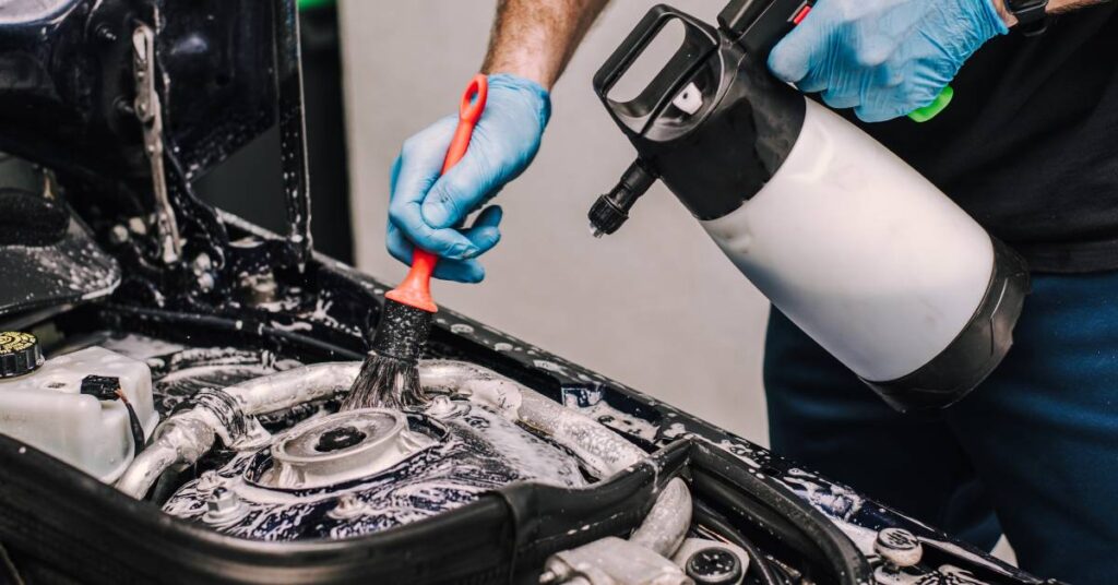 A close-up of a man spraying solvent on a car engine and cleaning it with an orange brush. Soap suds are on the engine.