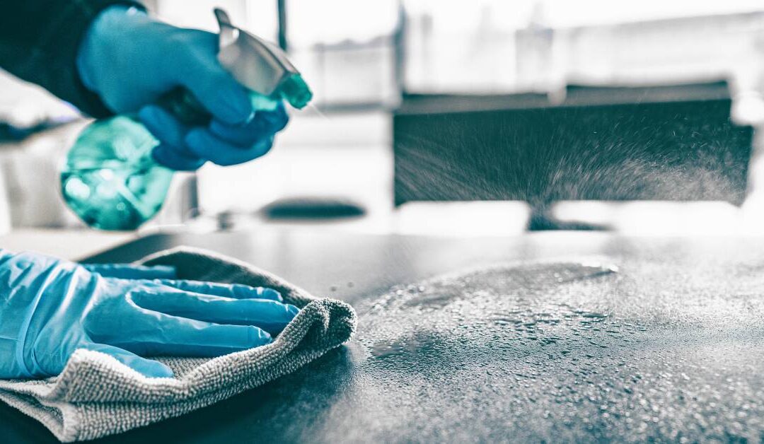 A close-up of someone spraying cleaner on a table and wiping it with a gray towel. The person is wearing blue gloves.