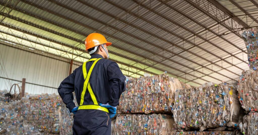 A worker stands in a large warehouse with multiple stacks of recycled materials. He's wearing protective gear.