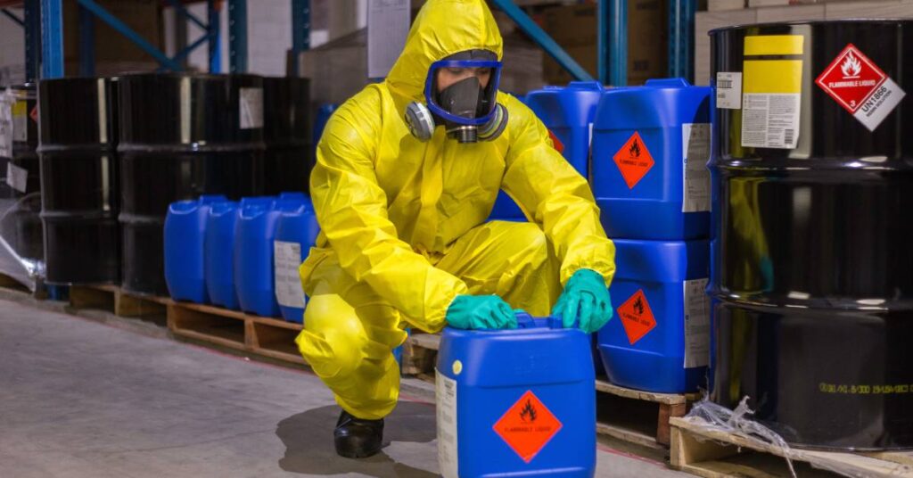 A person in a yellow protective suit with a ventilator mask kneels to open a blue chemical container.