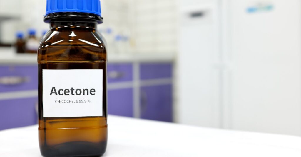 A dark bottle of acetone on a white table. The bottle says "Acetone" with the solvent's chemical formula.