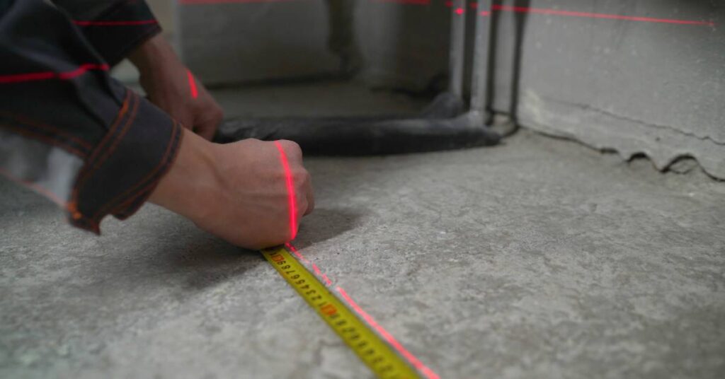 Close-up of a man measuring a dark concrete floor. He uses a standard tape measurer and a laser measurer.