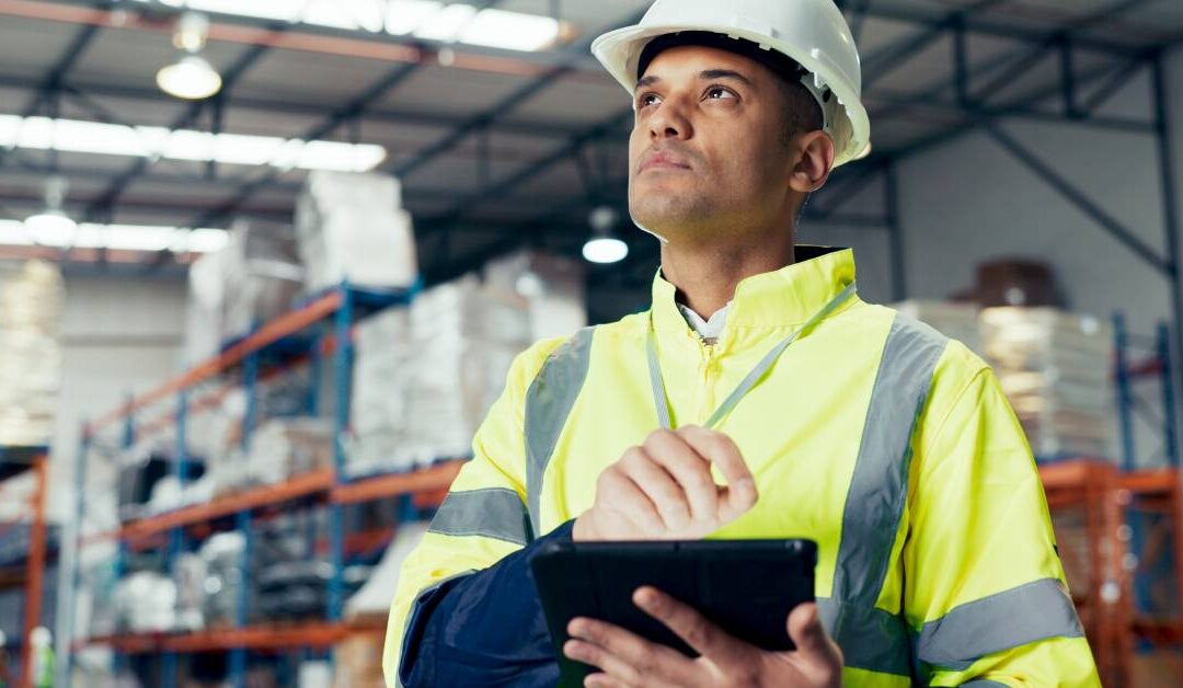 A worker stands in a warehouse wearing a reflective jacket and hard hat. He holds a tablet and looks up.