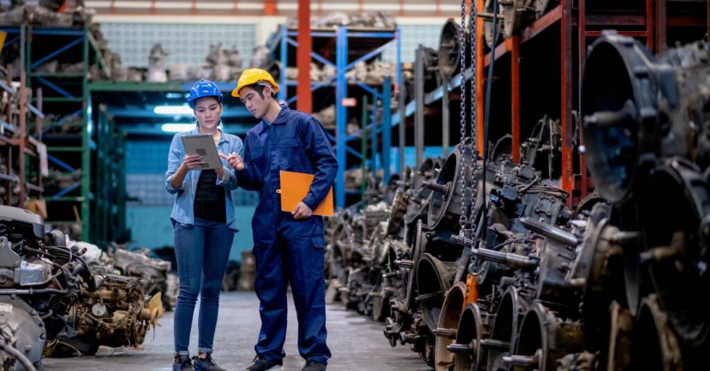 Two workers in an automotive manufacturing plant. One worker holds a tablet, and the other points to the screen.