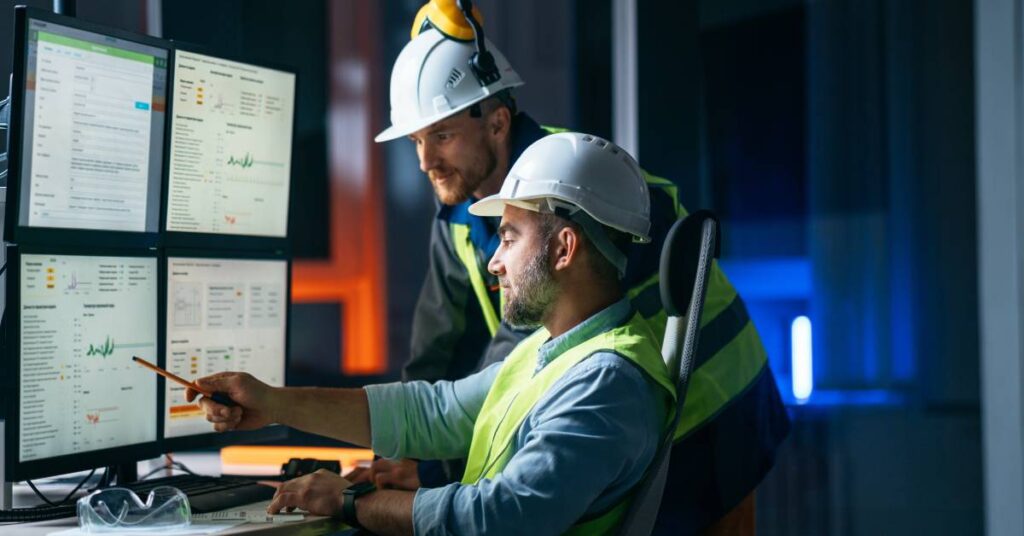 Two workers near multiple computer screens read data. The worker sitting down points at a graph with a pencil.