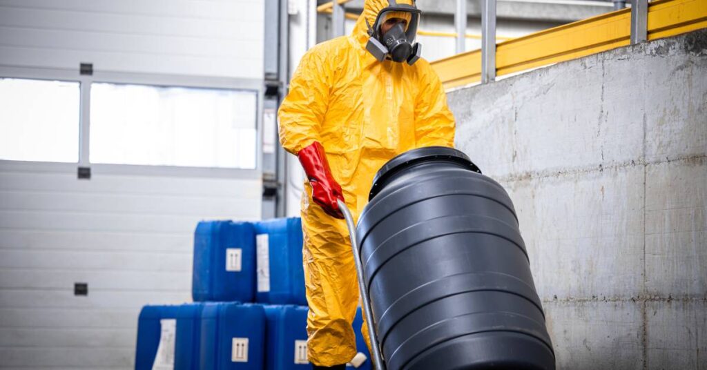 A worker in protective gear wheels a large, black solvent barrel inside a facility. Blue solvent containers sit in the back.