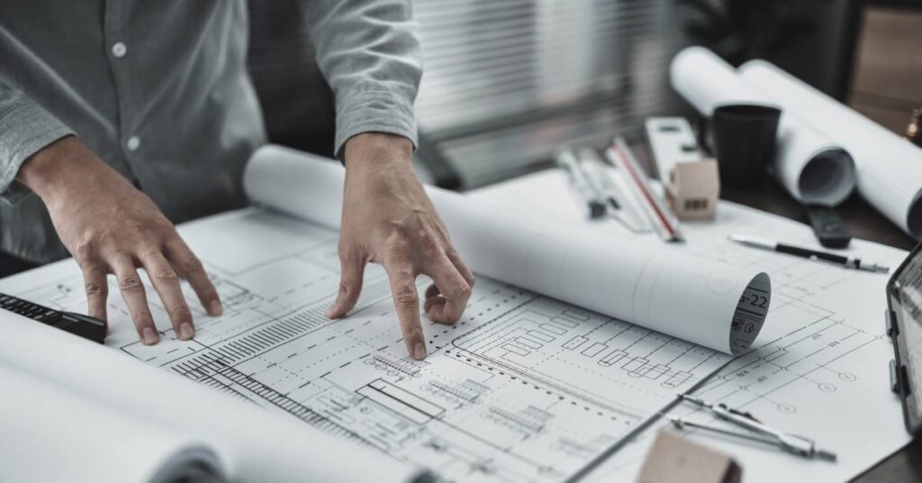 A close-up of hands holding open factory blueprints on a table. Other rolled blueprints and more items are nearby.