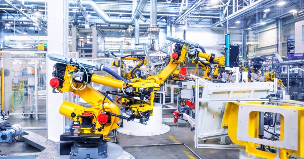 Robotic arms in a manufacturing plant assembly line. The arms are yellow with red and black components.