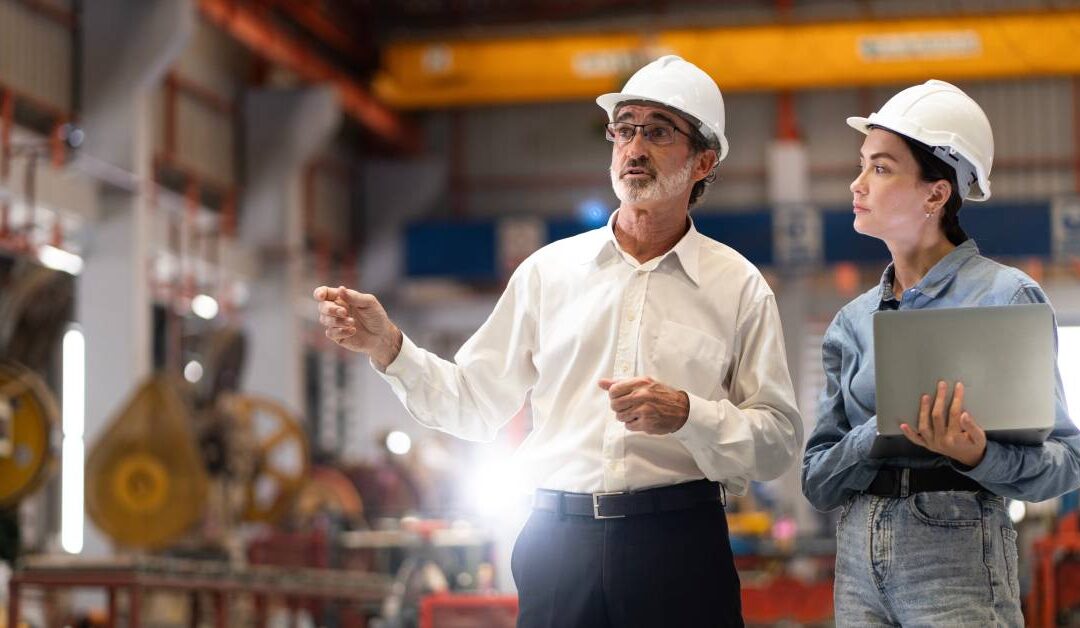 Two workers wear business attire and stand in a manufacturing plant. Both workers have hard hats, and one holds a laptop.