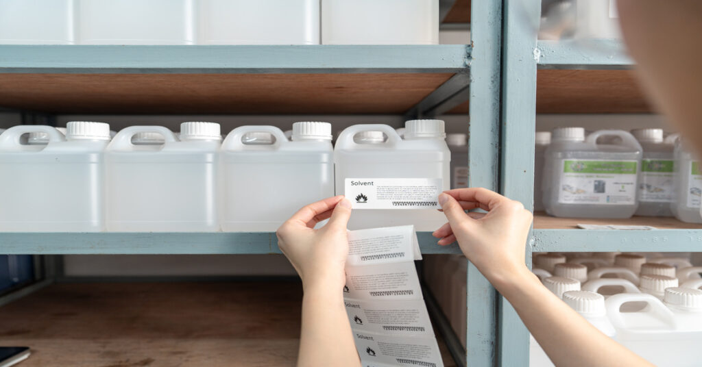 A woman placing solvent label on plastic chemical bottle on warehouse shelf with identical containers.