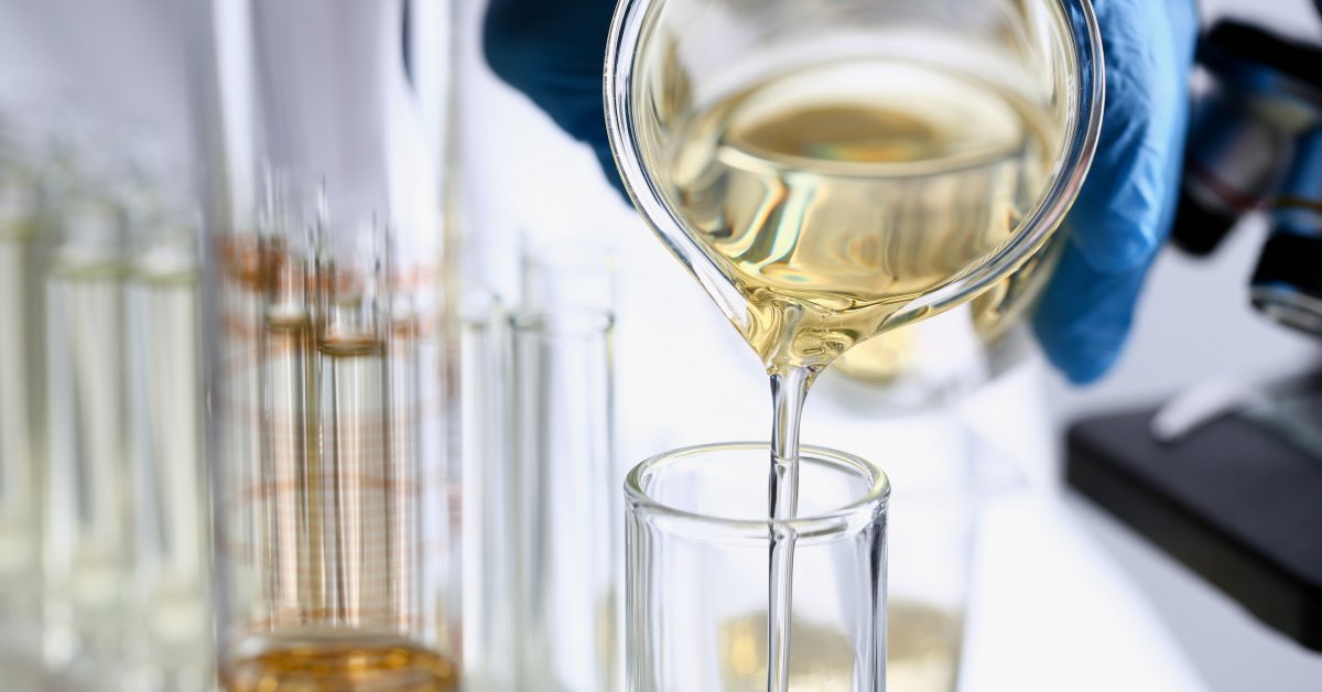 A person wearing blue gloves is holding a beaker with an orange liquid while standing beside a counter and a microscope.