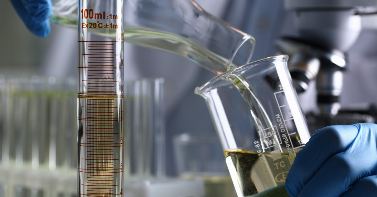 A person is pouring a clear liquid from one beaker into another while standing near a graduated cylinder and a microscope.