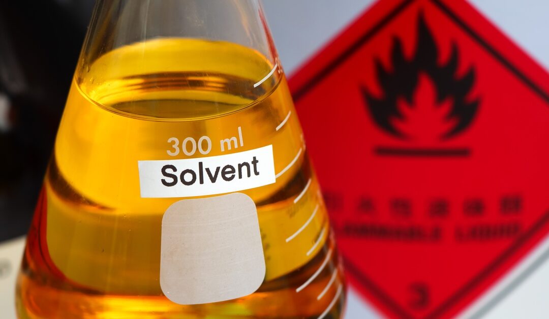 A glass beaker filled with yellow solvent sitting on a lab bench in front of a bold flammable warning label.