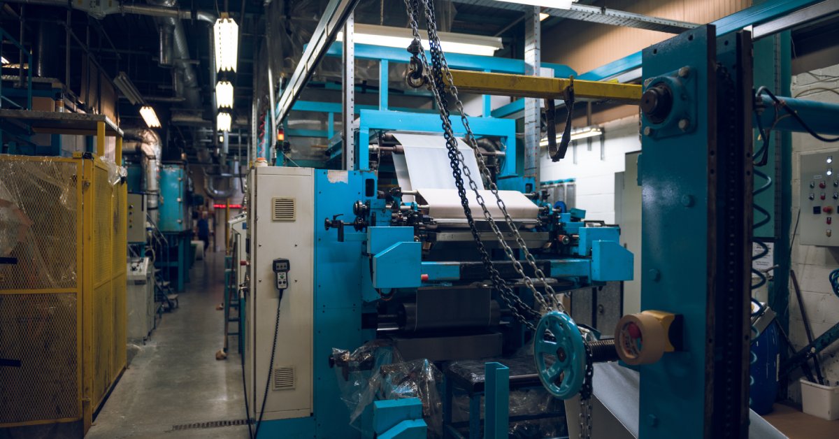 Blue automated machinery with rollers and metal arms sitting in a fluorescent-lit production room.