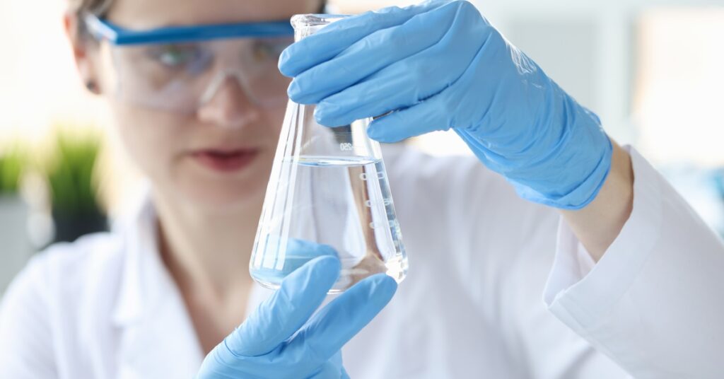 Woman wearing safety goggles, a lab coat, and blue gloves holding a clear Erlenmeyer flask containing a transparent liquid.