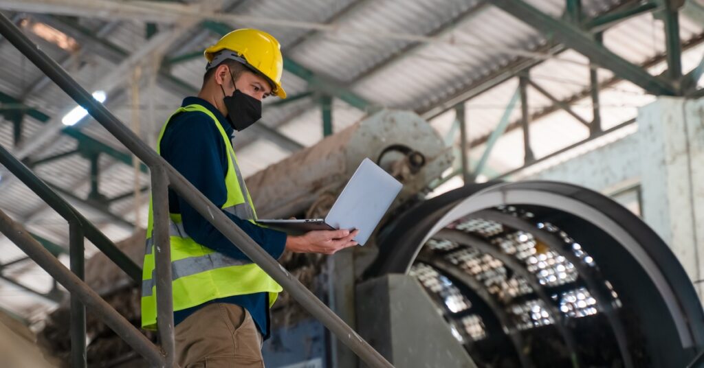 A worker in a hard hat and safety vest using a laptop while standing on metal stairs inside a large factory building.