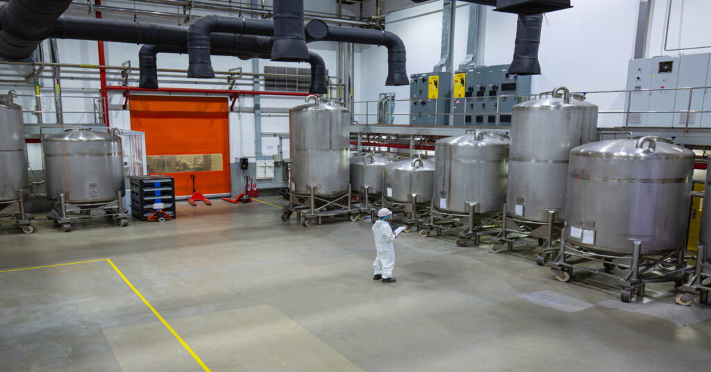A person in white safety gear standing near stainless steel tanks inside a large chemical processing facility.