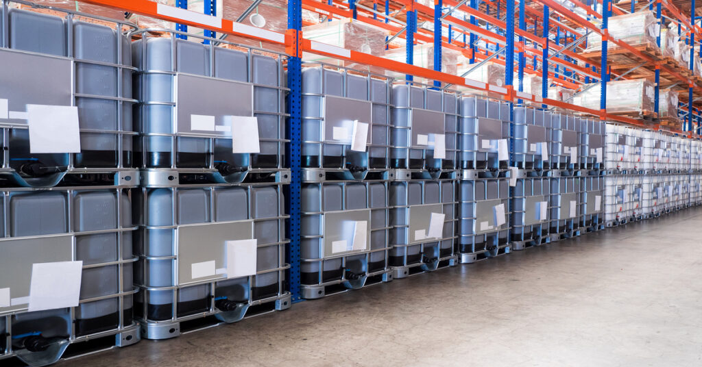 Rows of IBC liquid containers with white labels line the warehouse floor under bright industrial lighting.