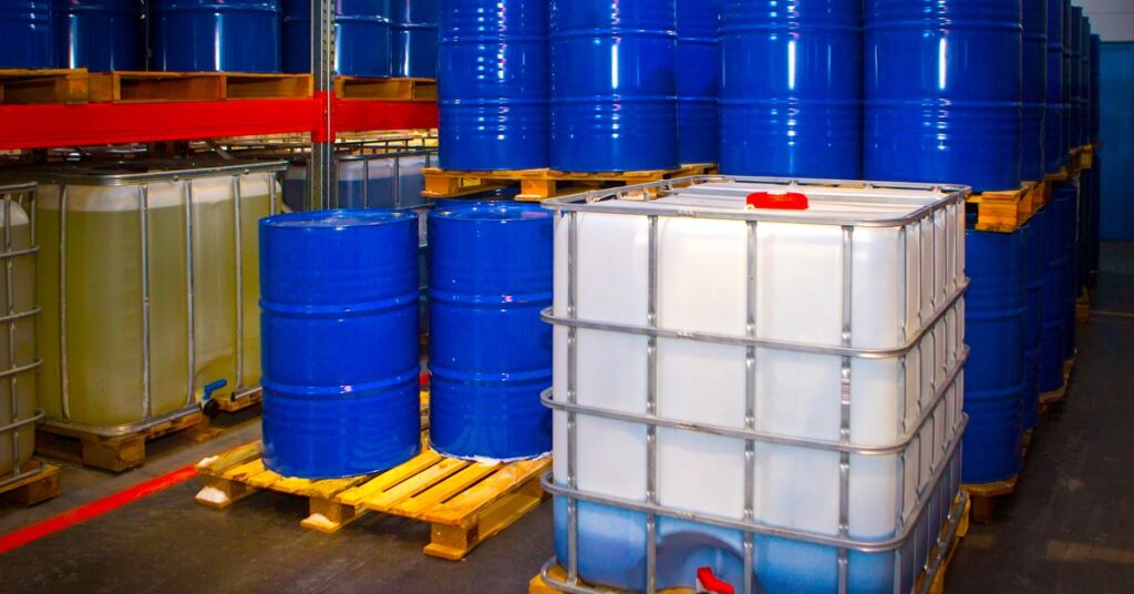 Dozens of blue metal drums and white IBC containers stacked on wooden pallets inside a chemical warehouse.