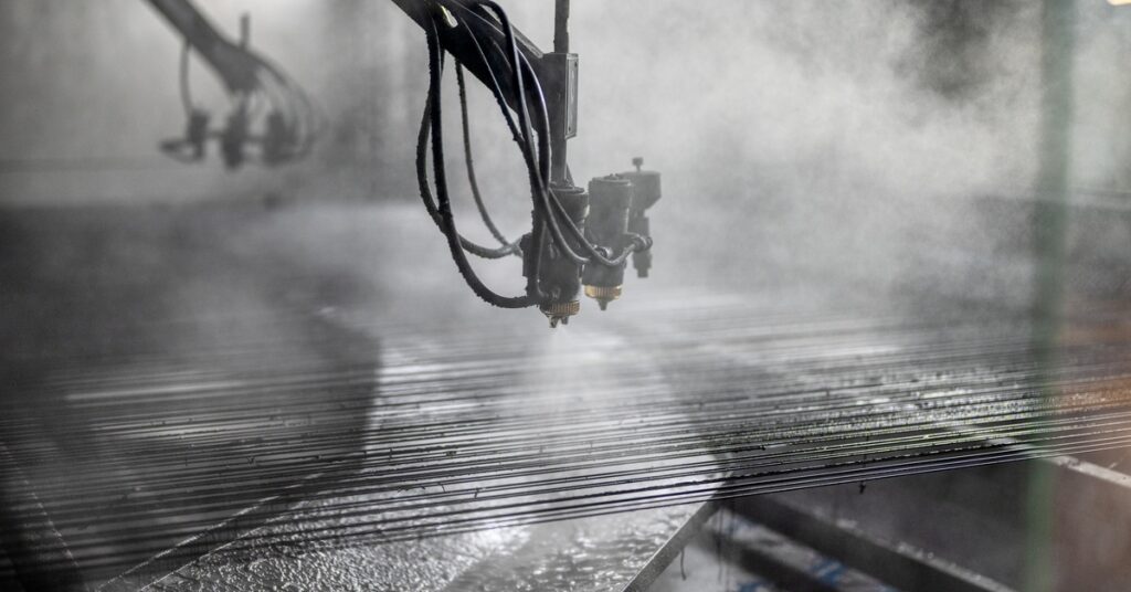 An industrial spray coating machine releasing fine mist onto metal rods inside a manufacturing chamber.