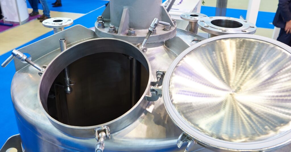 An open stainless steel paint dissolving tank with a circular lid and multiple valve attachments in a factory booth.