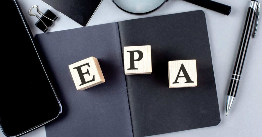 Wooden blocks labeled E, P, and A are sitting on a black notebook surrounded by a magnifying glass, pen, phone, and binder clip.
