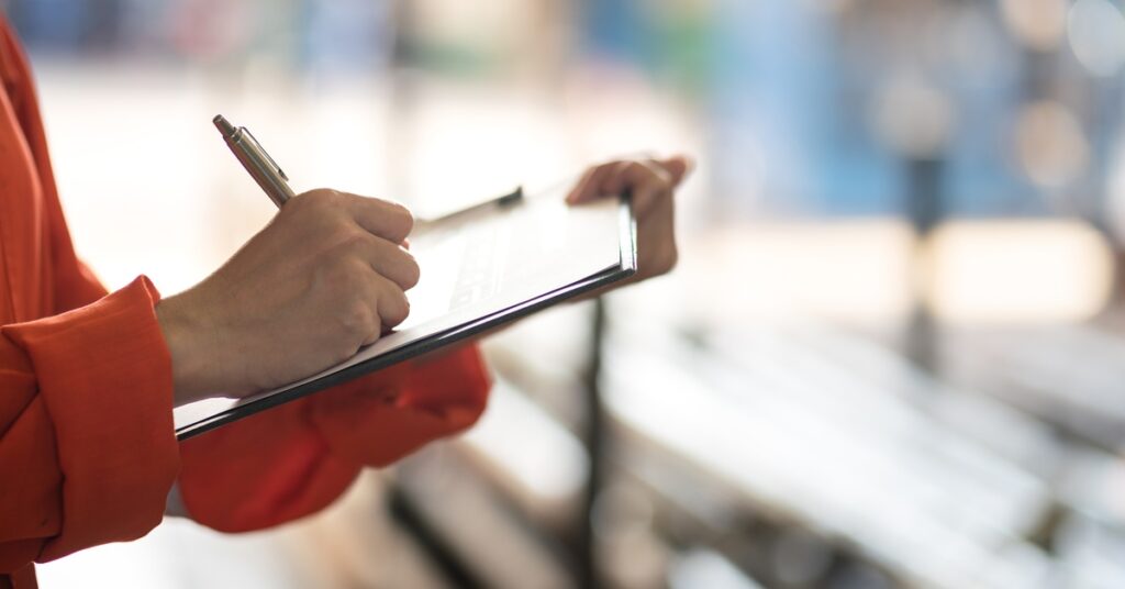 A person in an orange uniform writing with a pen on a clipboard while standing in a large indoor industrial space.