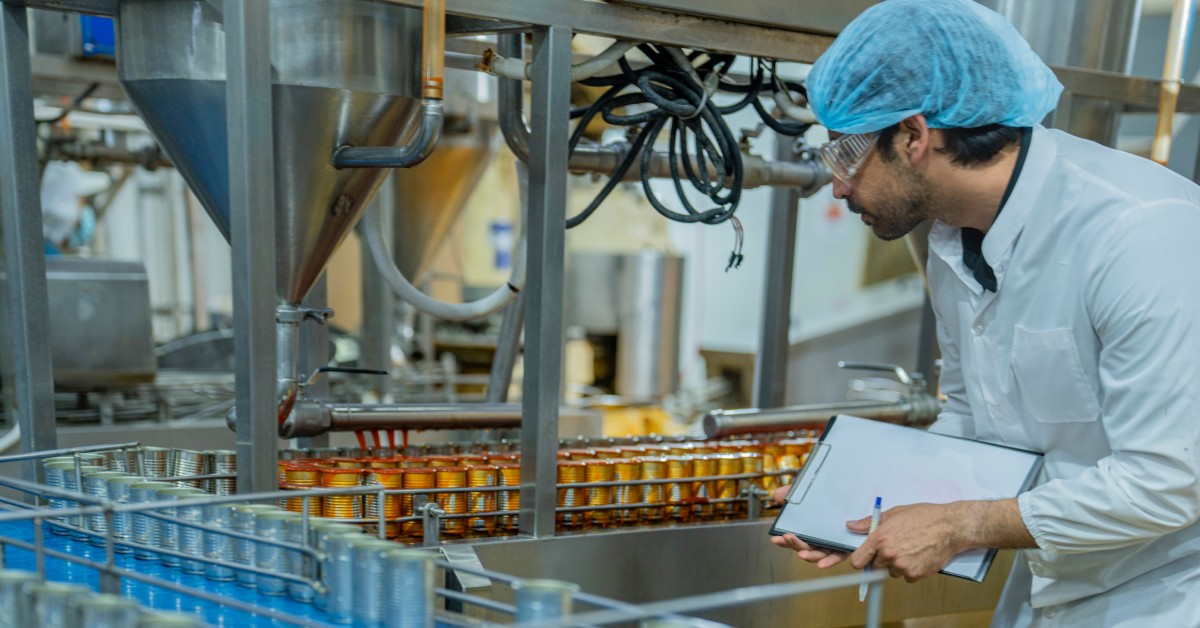 A man in a lab coat and hair net holding a clipboard while observing rows of cans under metal equipment in a food factory.