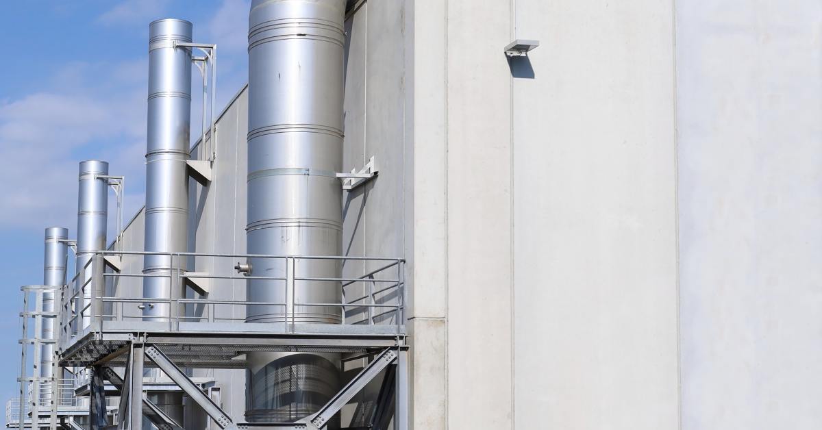 Silver industrial ventilation ducts and pipes attached to the side of a white building against a clear blue sky background.