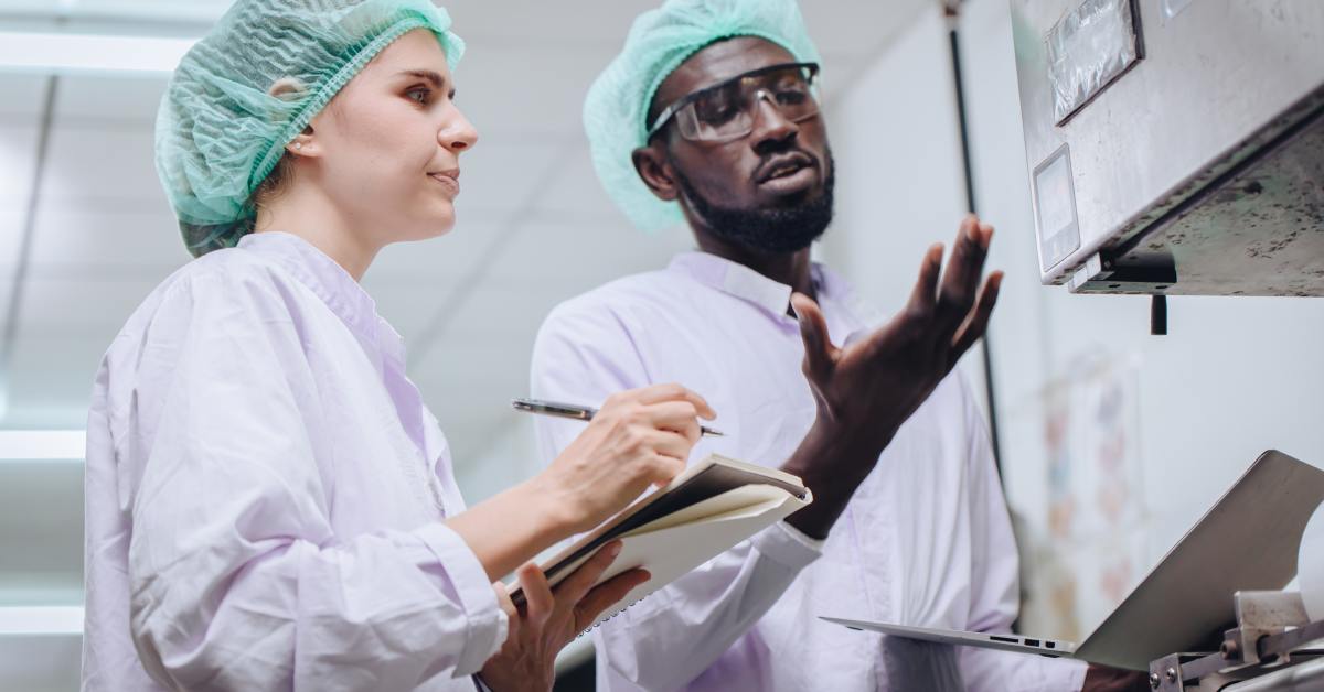 Two people in lab coats and hairnets standing by machinery. One is writing in a notebook while the other gestures with one hand.