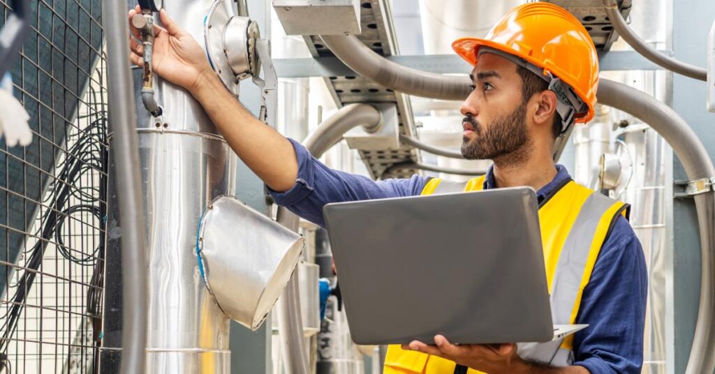 A person in an orange hard hat and safety vest using a laptop while inspecting industrial pipes and gauges inside a facility.