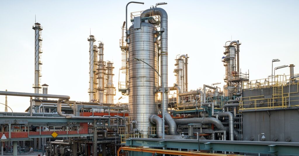 A large factory contains several metal distillation columns and pipes. A blue sky appears behind the factory.