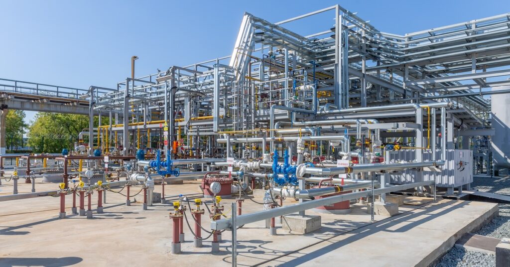 Several silver metal pipes come out of the side of a white factory. There are blue shut-off valves around the pipes.