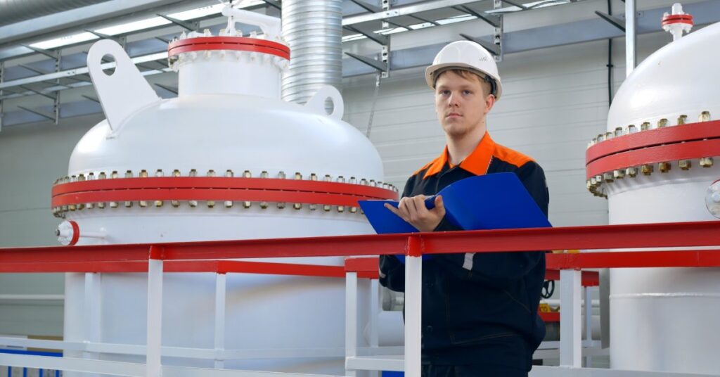 Man in white hard hat and dark uniform holds blue clipboard while standing near large white industrial tanks with red bands.