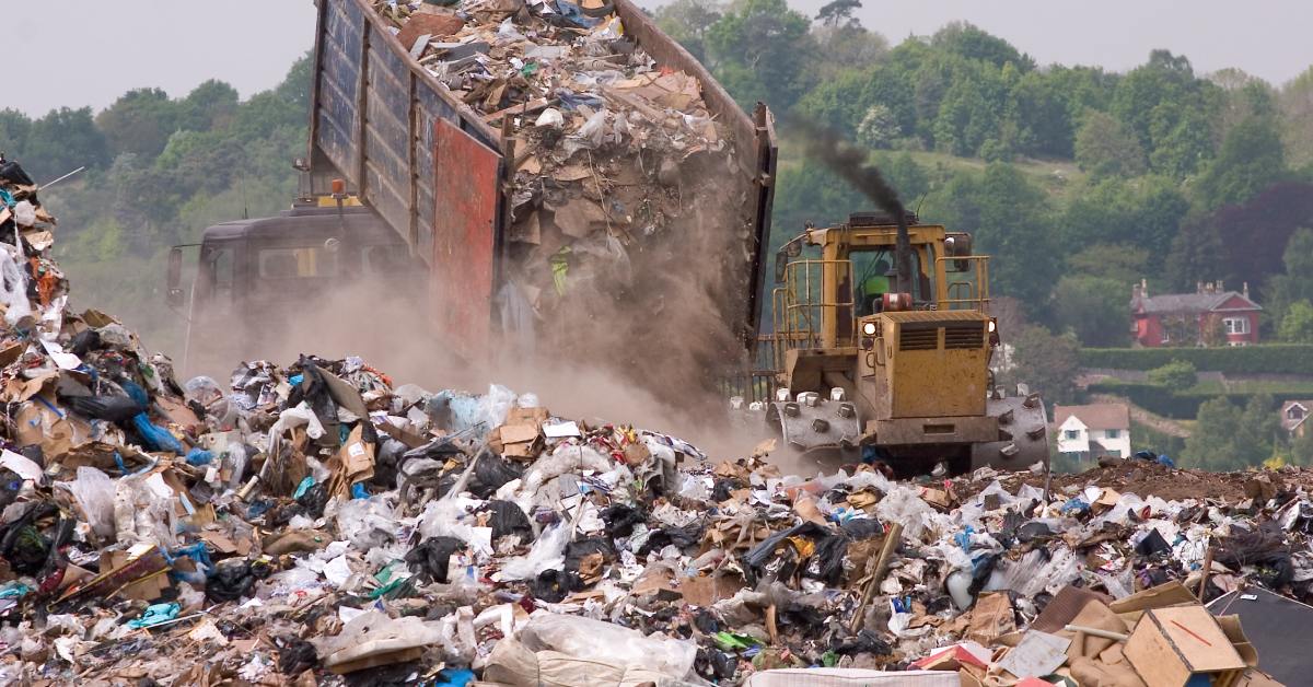 A dump truck is dropping a load of waste onto a landfill while a bulldozer pushes the waste to flatten it out.