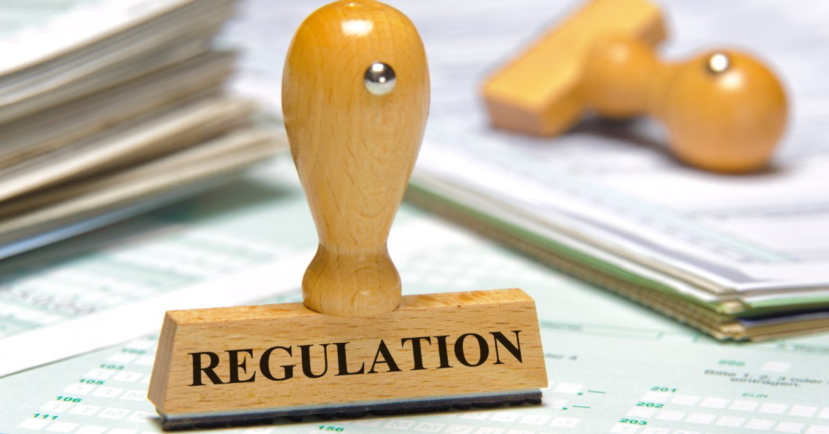 A close-up view shows a wooden stamp with a word "regulation" in black ink across it. It sits on a pile of papers.