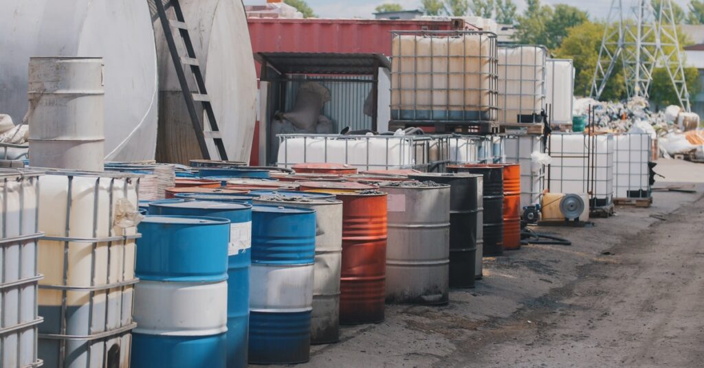 A mixture of colorful metal barrels and square plastic storage containers surrounded by metal cages sit outside a facility.