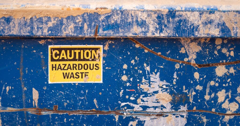 A blue dumpster with white and tan paint streaks features a yellow sticker that reads "Caution Hazardous Waste."