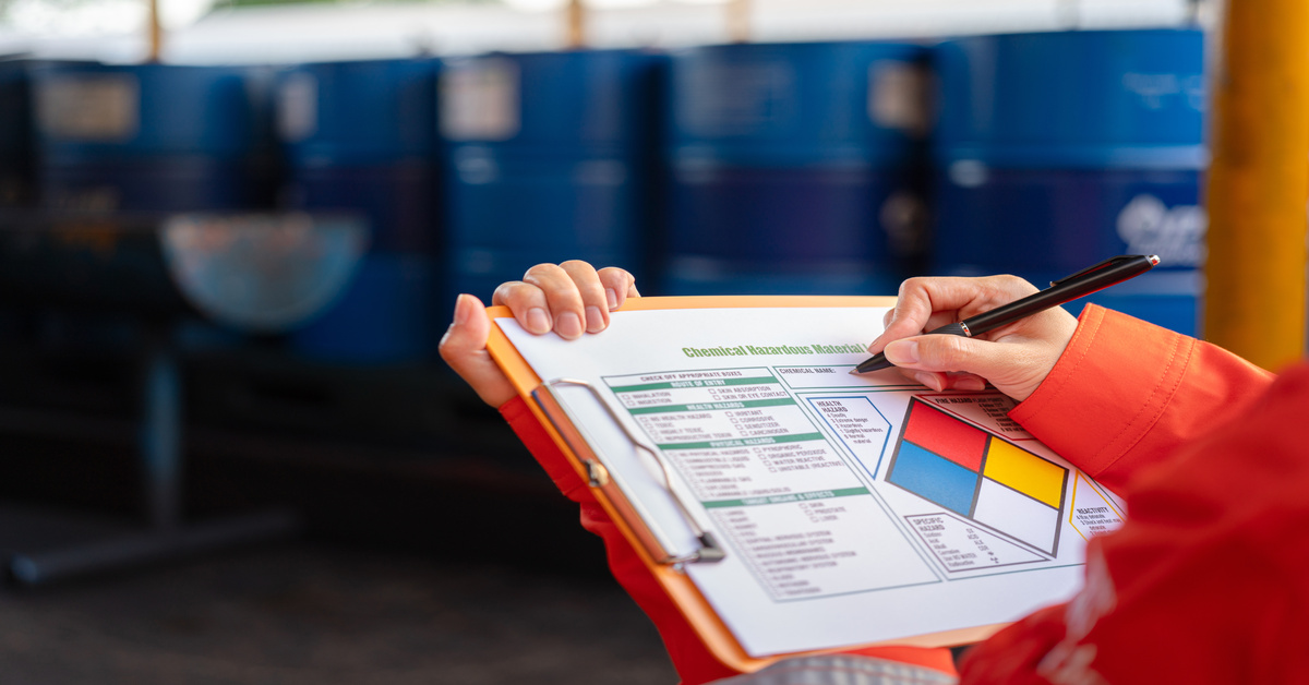 A close-up view shows a safety checklist sitting on a red clipboard. A person uses a black pen to write on the checklist.